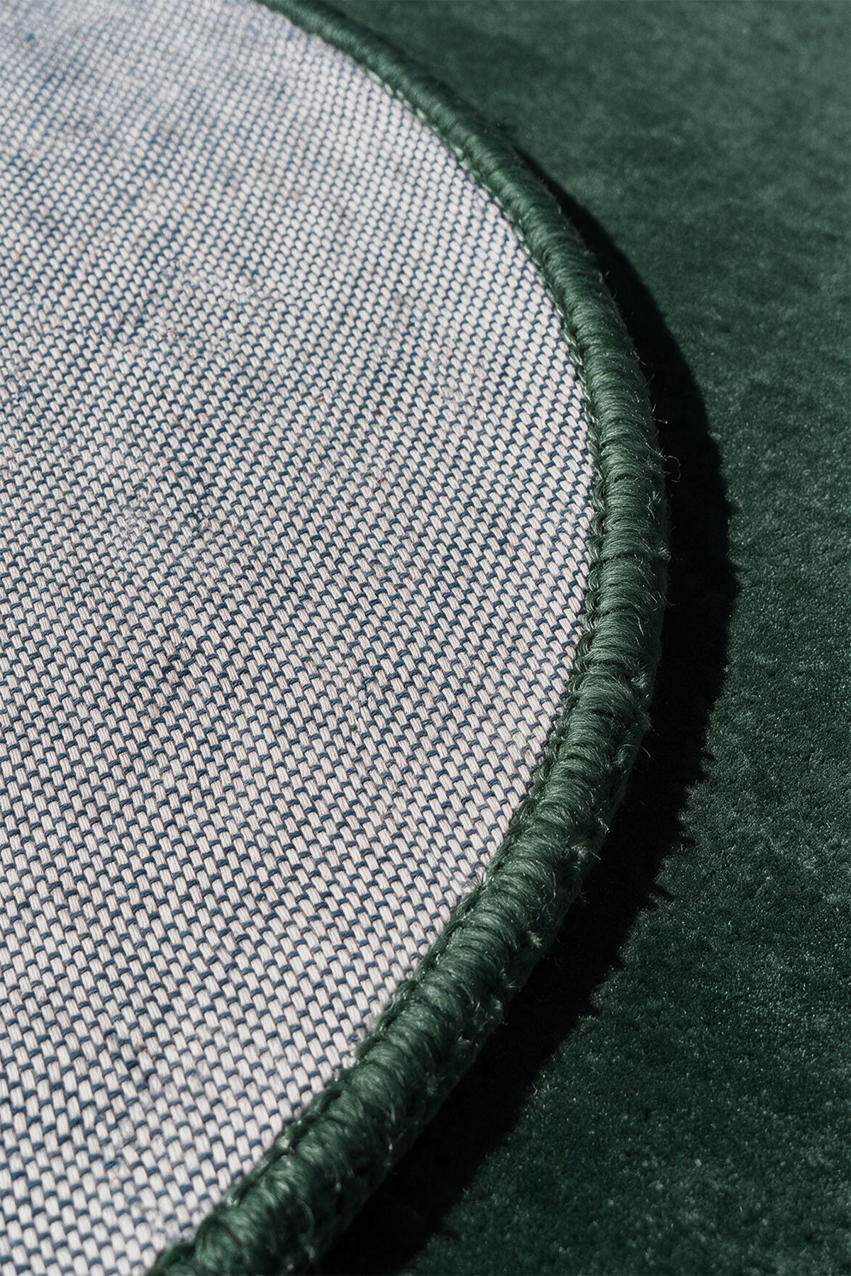 Close-up detail of a round rug showing a woven light gray fabric surface, a dark green stitched border, and a dark forest green velvet-like background | Cotton backing