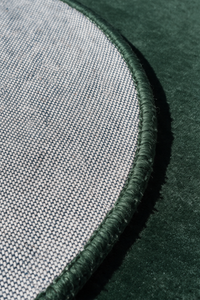 Close-up detail of a round rug showing a woven light gray fabric surface, a dark green stitched border, and a dark forest green velvet-like background | Cotton backing