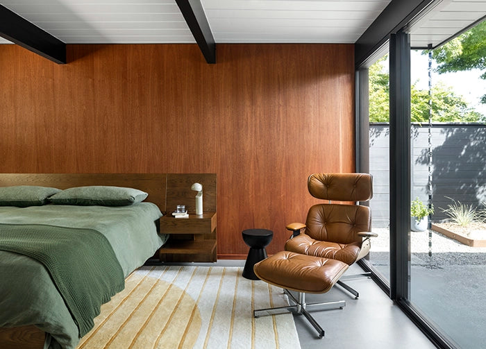 A mid century modern bedroom interior with a bed dressed in olive green bedding, a wood-paneled wall, gold geometric rug, a brown leather lounge chair with matching ottoman, and large floor-to-ceiling glass doors opening to an outdoor patio.