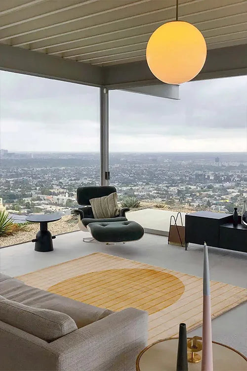 Modern living room with a view of a cityscape, featuring a round pendant light, gold contemporary rug and comfortable furniture.