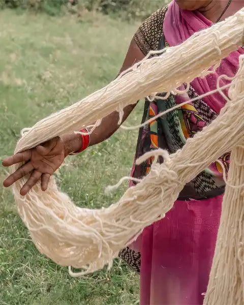 A person outdoors holding up a large bundle of off-white wool yarn over a grassy field. The person is wearing a pink and black patterned garment and a red wristband.