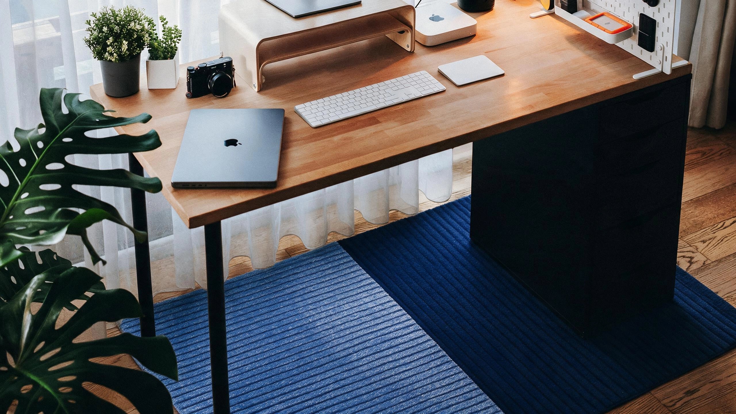 A modern wooden desk with a black pedestal base, styled with a laptop, keyboard, and desk accessories, set on a blue striped rug in a bright room.