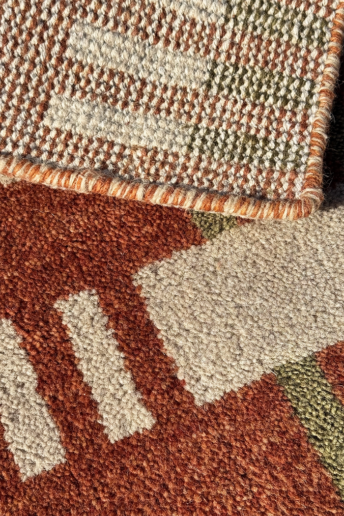 Close-up of a textured area rug with a geometric pattern in rust brown, cream, and olive multicolor, showing the pile texture and corner detail.