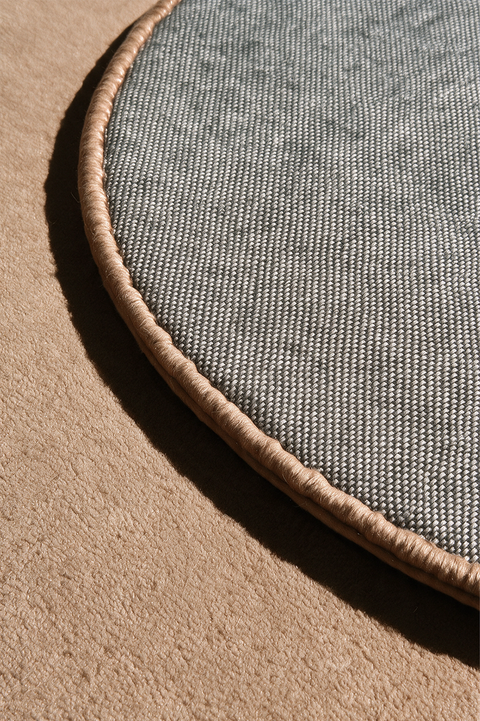 Close-up of a round rug showing its textured gray woven underside and beige stitched edge, light brown wool | Cotton backing