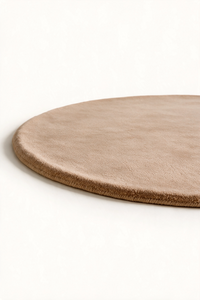 A close-up of a round, low-profile rug in a warm taupe or light brown, shown on a clean off-white background.
