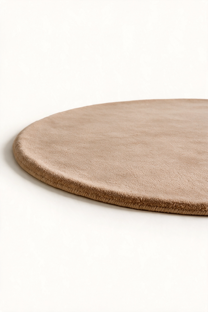 A close-up of a round, low-profile rug in a warm taupe or light brown, shown on a clean off-white background.