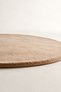 A close-up of a round beige light brown area rug with a soft, low-pile texture, shown resting on a neutral light gray surface.