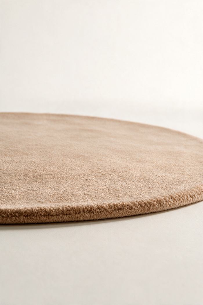 A close-up of a round beige light brown area rug with a soft, low-pile texture, shown resting on a neutral light gray surface.