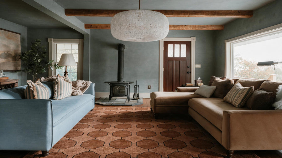 A cozy living room with a light blue sofa on the left, a beige sectional sofa on the right, a geometric brown rug, a black wood-burning stove, and a large white textured pendant light hanging from exposed wooden beams.