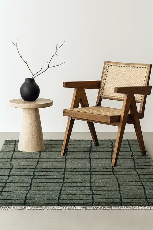 Wooden armchair with woven backrest next to a small round table with a black vase on a green wool rug.