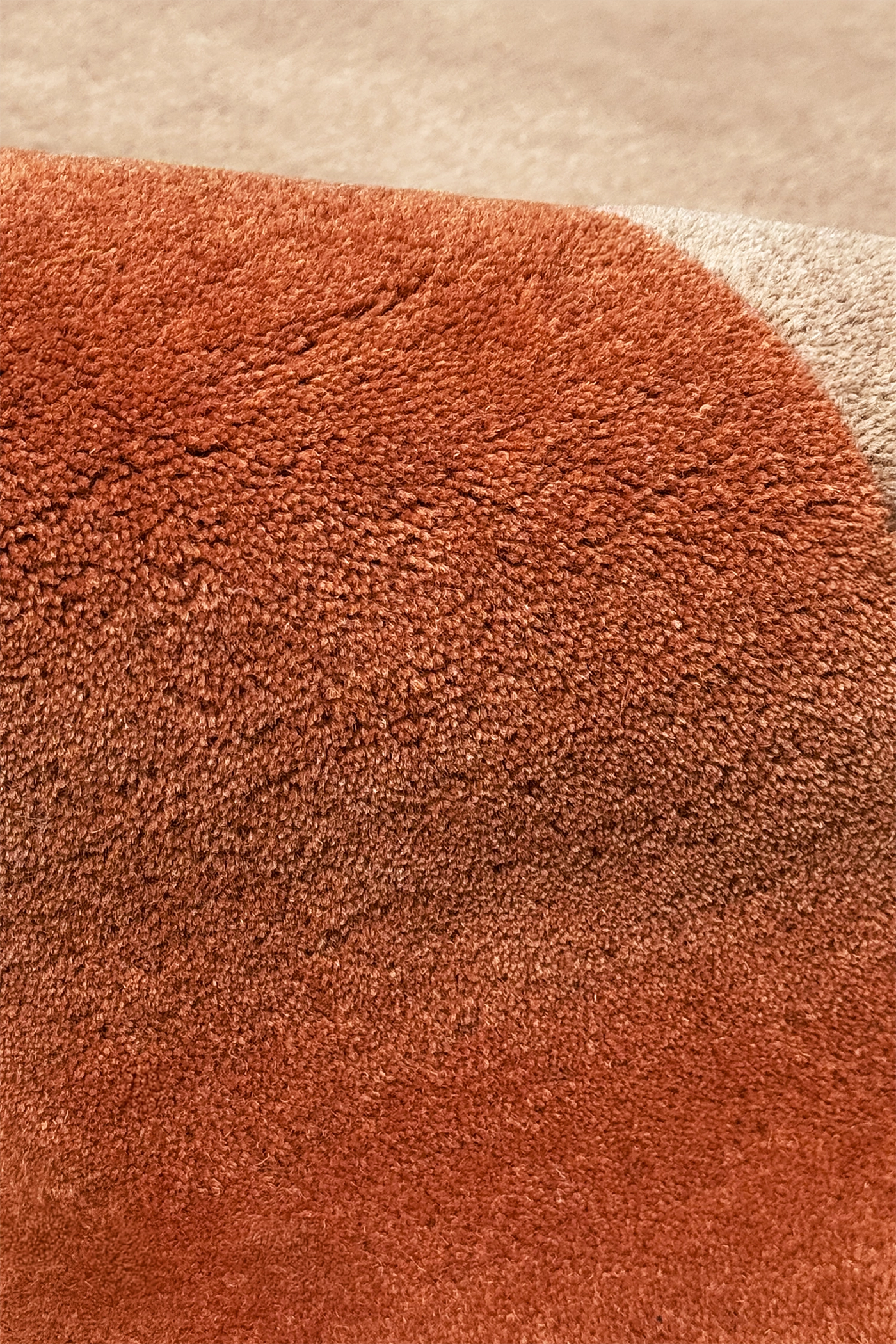 Close-up of a plush, high-pile rug in a warm rust or terracotta color, showing the dense loop texture and a curved seam leading to a lighter beige section.