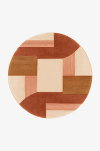 A round area rug with a geometric patchwork design in terracotta, dusty pink, beige, and brown, shown from a top-down view on a light gray background.
