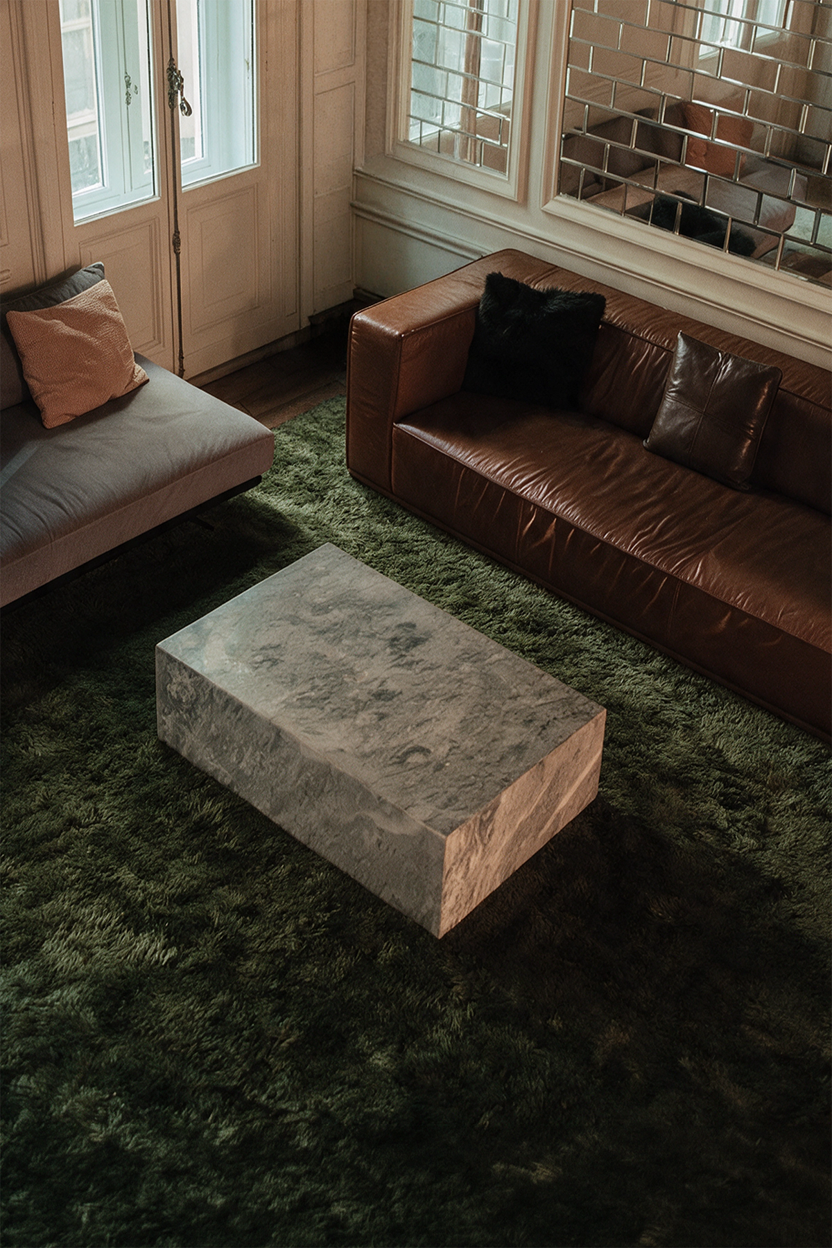 A modern rectangular coffee table made of textured gray stone sits on a dark olive green shag rug in a stylishly decorated living room with a brown leather sofa and a gray chaise lounge lifestyle