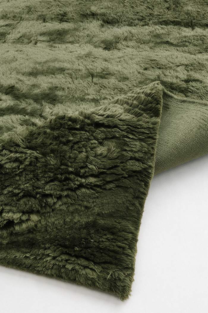 A dark olive green shaggy wool rug with a textured looped pile, folded over to show its soft underside on a clean white background.