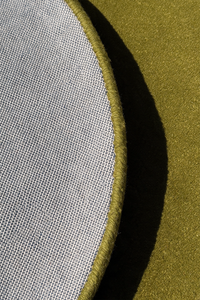 Close-up detail of a circular rug showing a light gray woven fabric edge with a thick olive green stitched border against a matching olive green textured background.