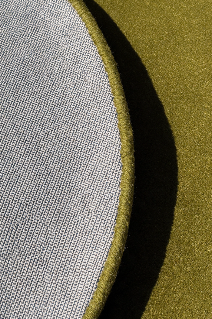 Close-up detail of a circular rug showing a light gray woven fabric edge with a thick olive green stitched border against a matching olive green textured background.