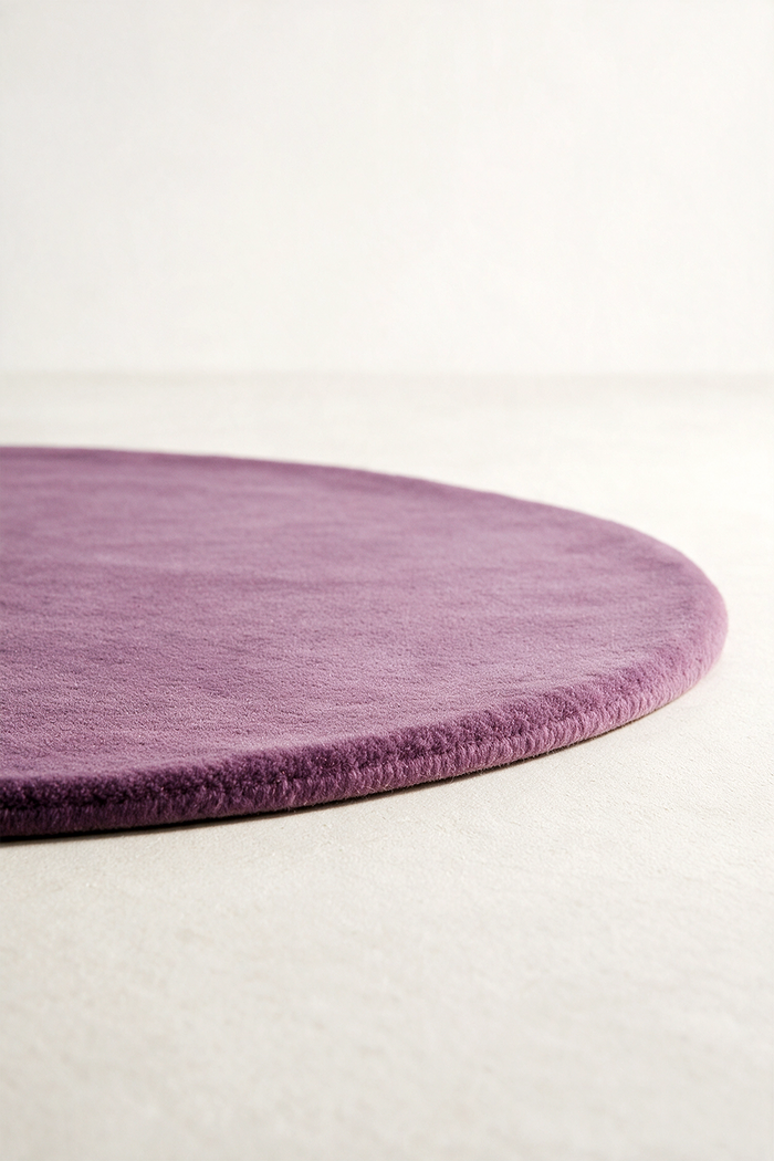 A close-up of a round plum purple rug with a short pile texture, shown on a light beige floor against a white background.