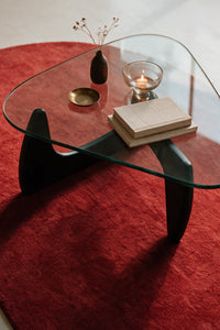 A modern low coffee table with a clear glass top and sculptural black metal legs, styled on a textured red rug with books, a candle, and a small vase.