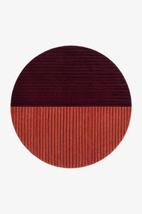 A round ribbed rug split horizontally into two colors: a dark burgundy top half and a terracotta orange bottom half, shown on a plain white background rust