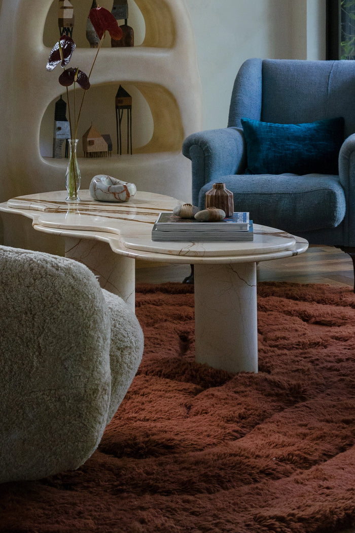 A styled interior scene featuring a sculptural white marble coffee table with organic wavy edges, set on a rust-brown shag rug. In the background, a blue armchair with a teal pillow and a cream-colored curved shelving unit are visible lifestyle