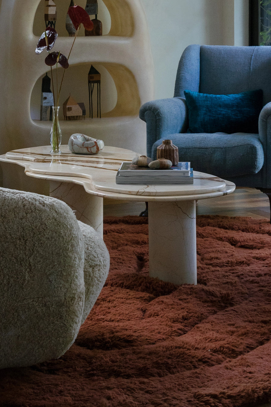 A styled interior scene featuring a sculptural white marble coffee table with organic wavy edges, set on a rust-brown shag rug. In the background, a blue armchair with a teal pillow and a cream-colored curved shelving unit are visible lifestyle