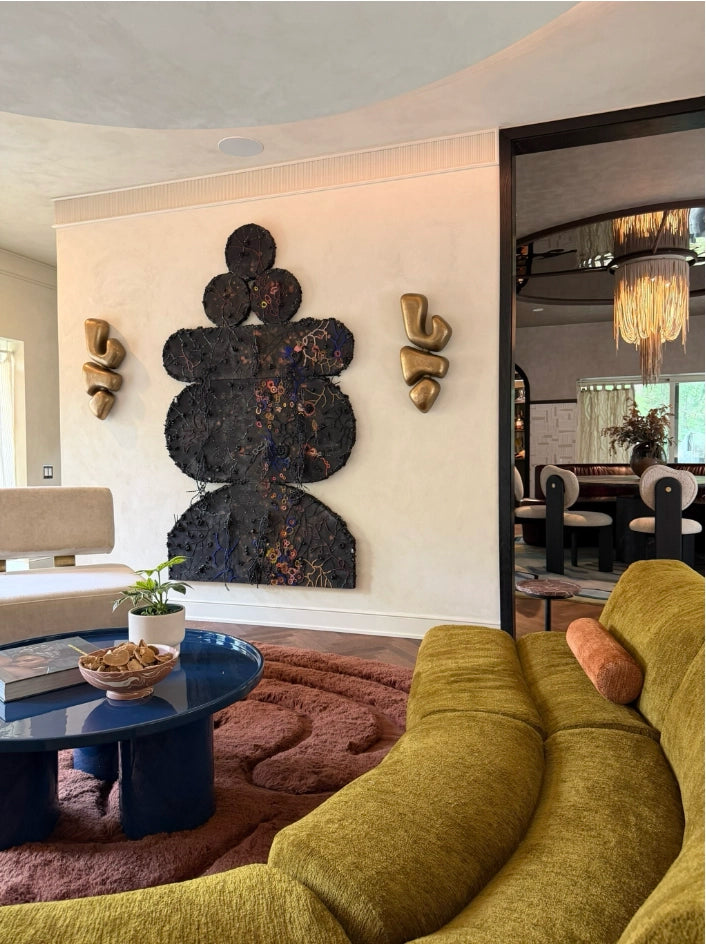 A modern living room interior featuring a large black abstract wall sculpture as the focal point, flanked by two gold abstract wall sculptures. In the foreground, a mustard yellow fabric sofa is partially visible, along with a blue coffee table and a textured rust-colored rug.