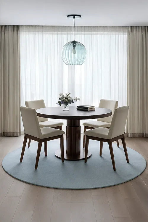 Dining room with a round wooden table and white chairs on a round baby blue wool rug, featuring a pendant light and large windows.