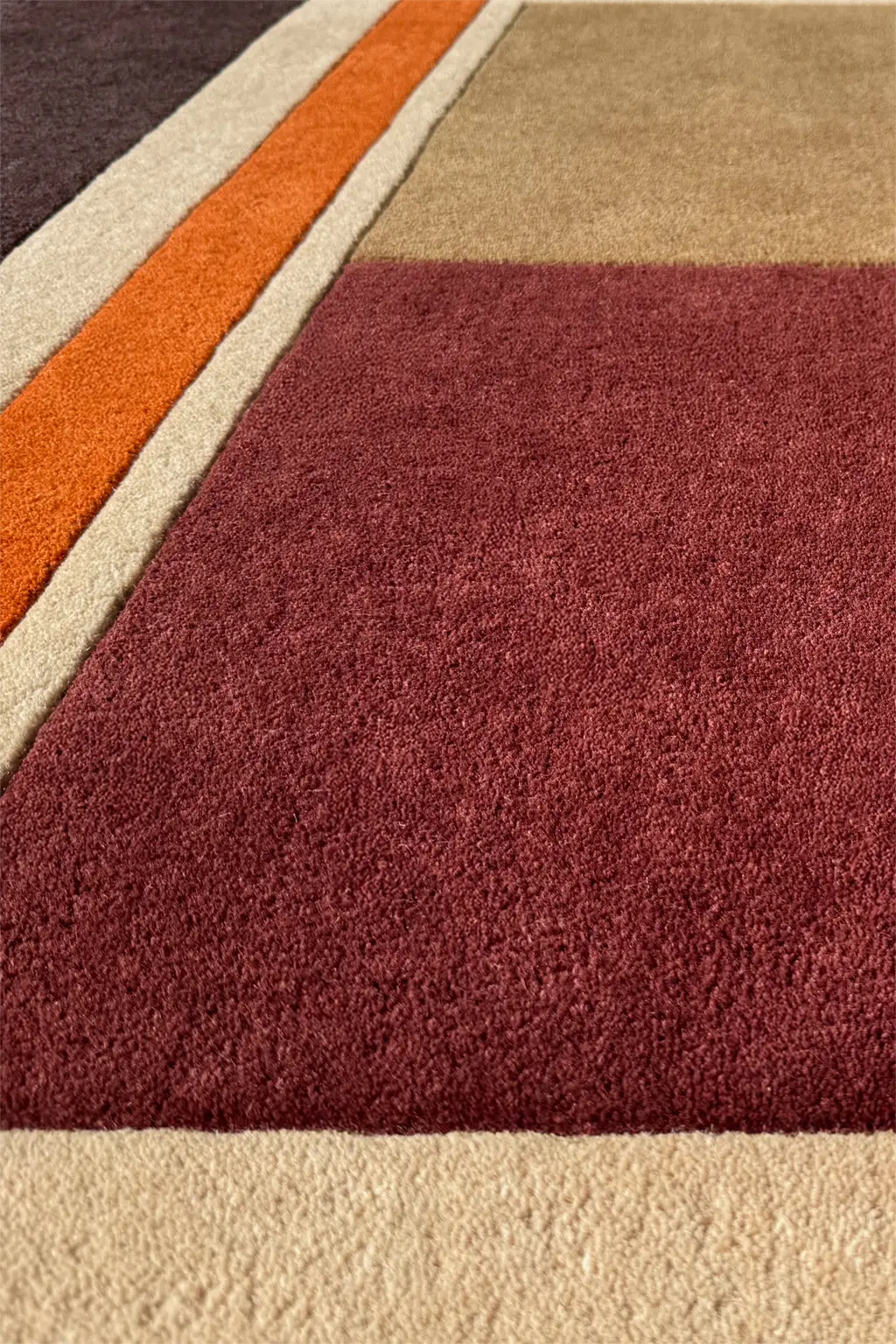 Close-up of a square area rug with a geometric color-blocked pattern featuring a large burgundy section, a beige section, and orange and cream stripes.