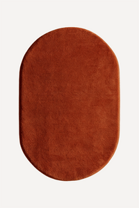An oval-shaped rug in burnt orange shown from a top-down view on a clean, off-white background.