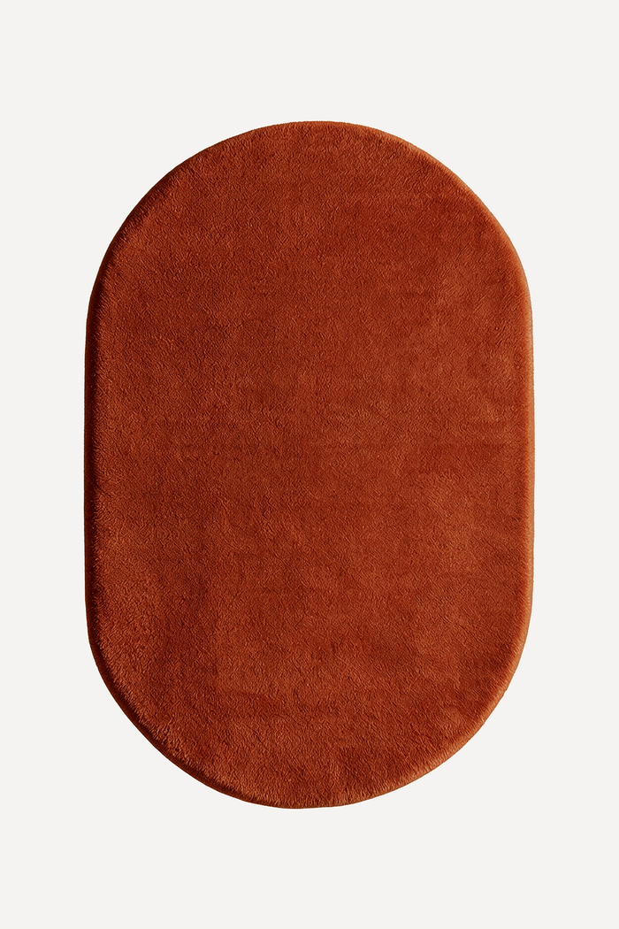 An oval-shaped rug in burnt orange shown from a top-down view on a clean, off-white background.
