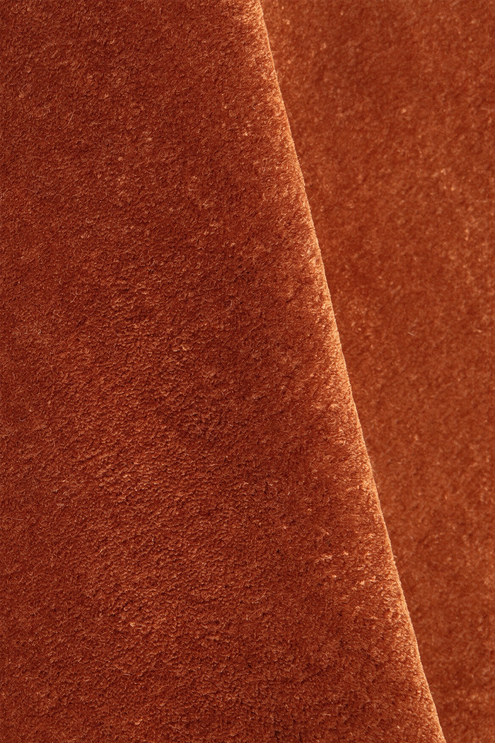 A close-up of a textured wool rug in a warm rusty burnt orange color, with a diagonal fold creating a soft shadow.