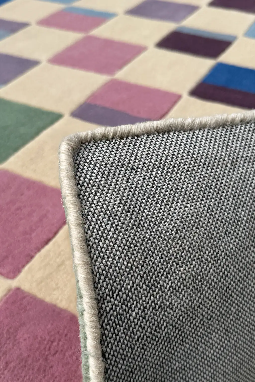 Close-up of a textured gray rug corner with a multicolored checkered pattern in the background.