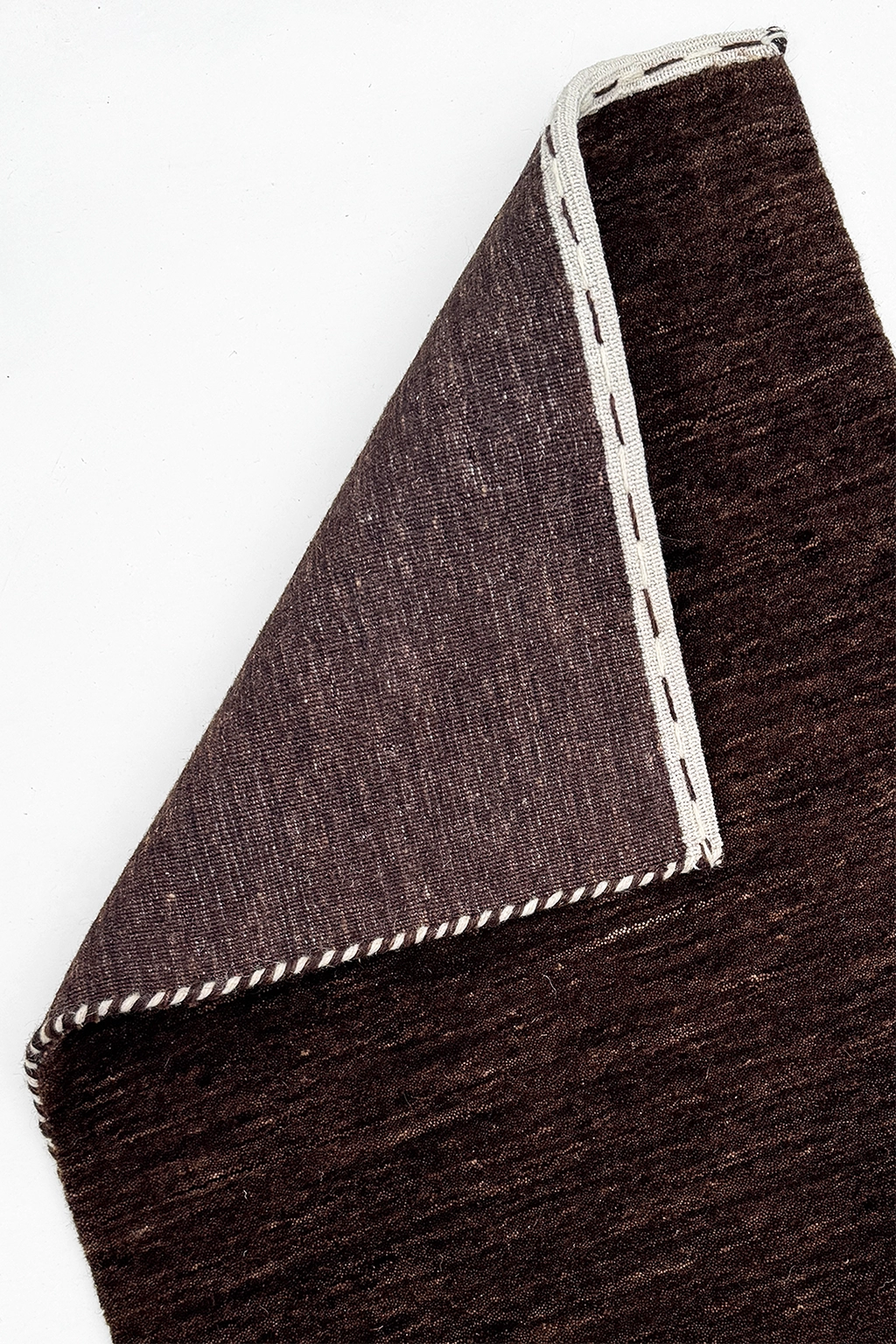 A close-up of a dark brown loomed runner rug corner, folded over to show the underside with white stitched edges, on a plain white background cocoa