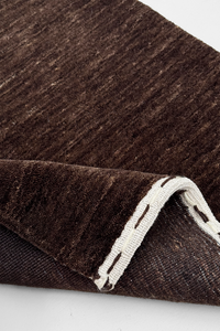 A close-up of a dark brown textured runner, likely wool or a wool blend, with a folded corner revealing a contrasting off-white stitched edge cocoa