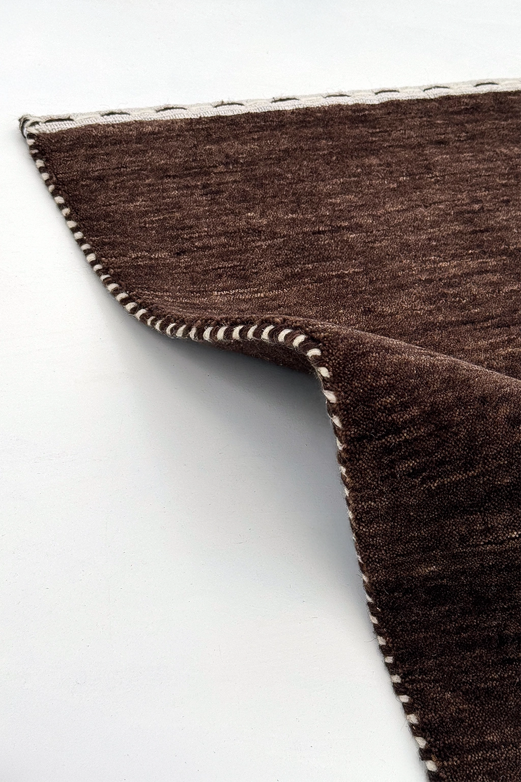 A close-up of a dark brown texture runner rug with a white whipstitched edge, folded over to show the underside and stitching detail against a plain white background cocoa