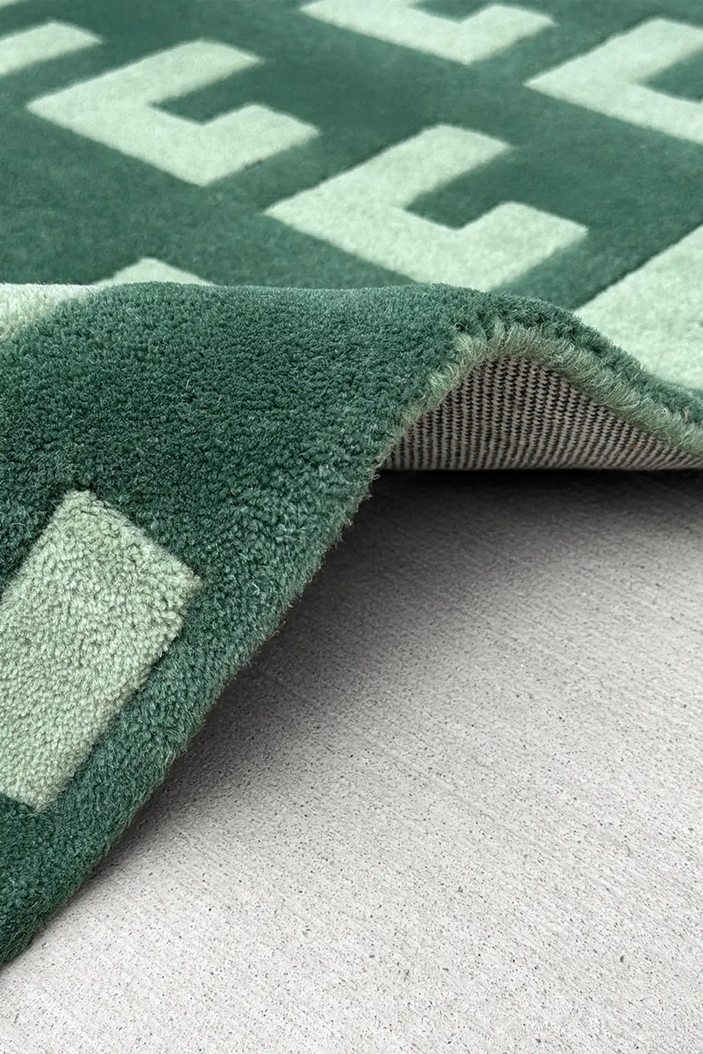 Close-up of a green textured square rug with geometric patterns on a gray surface.