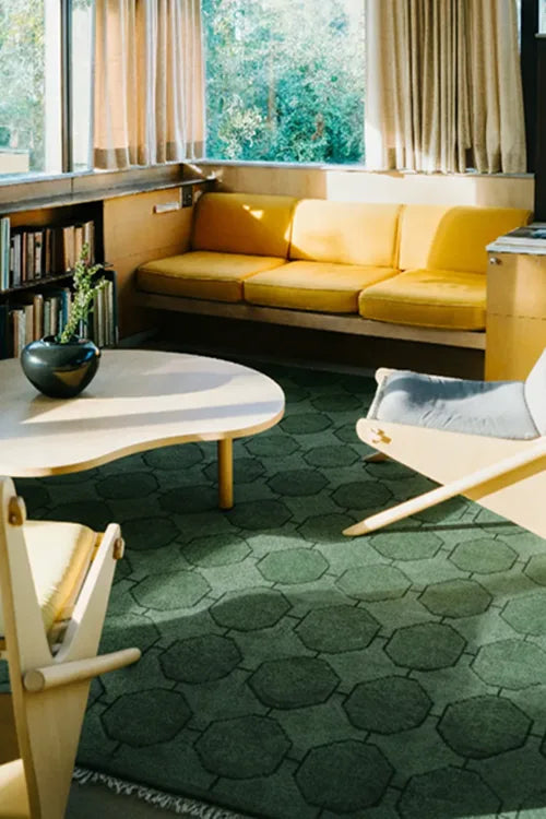 Modern living room with yellow sofa, green rug, and large windows.