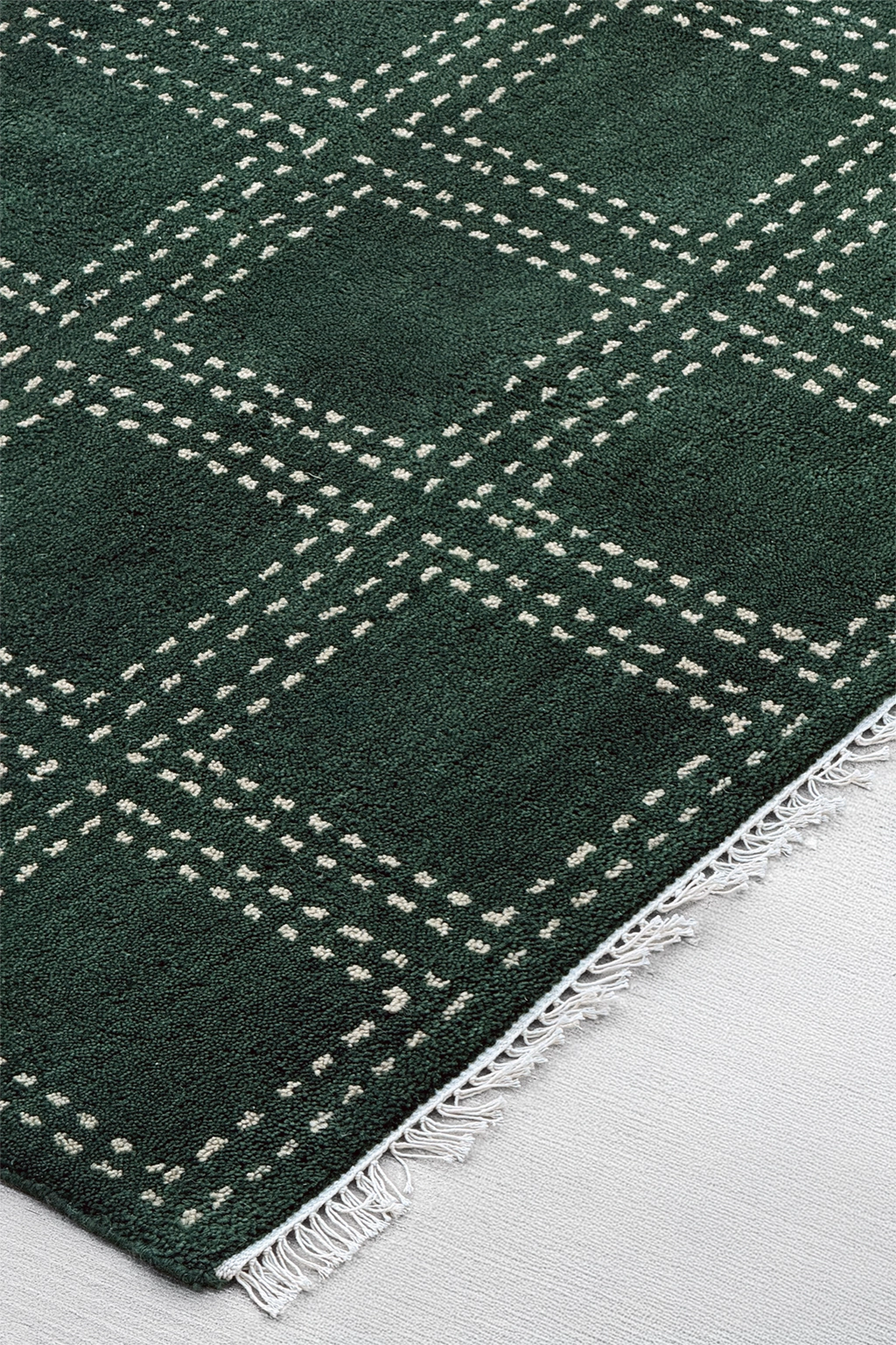 A close-up of a dark green square area rug with a white dotted plaid pattern and white fringe on a light gray floor.