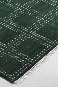 A close-up of a dark green square area rug with a white dotted plaid pattern and white fringe on a light gray floor.