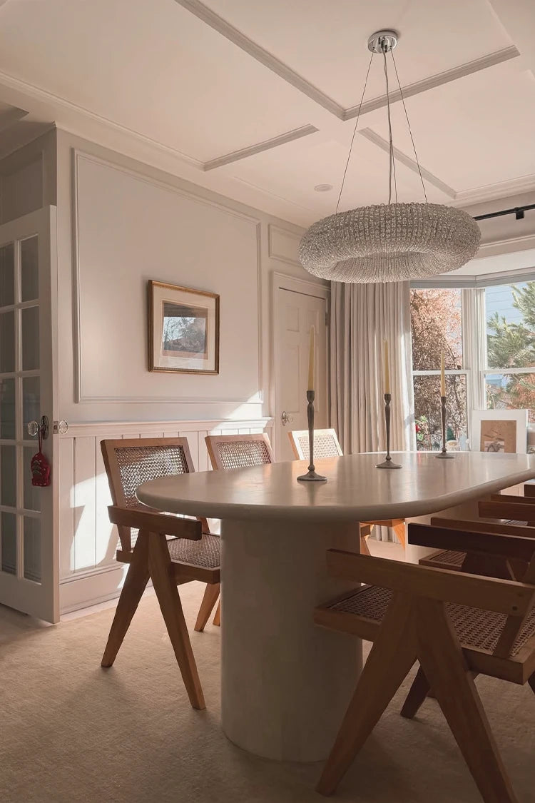 Dining room with a round table and wooden chairs, featuring a chandelier and large window.