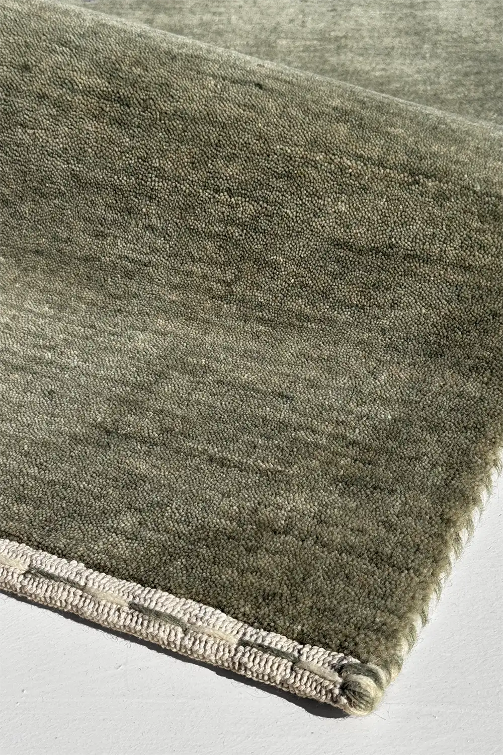 Close-up of a textured soft sage green wool rug with a white border on a light gray background