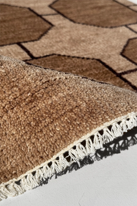 Close-up of a light brown geometric patterned rug with white fringe on a light gray floor.