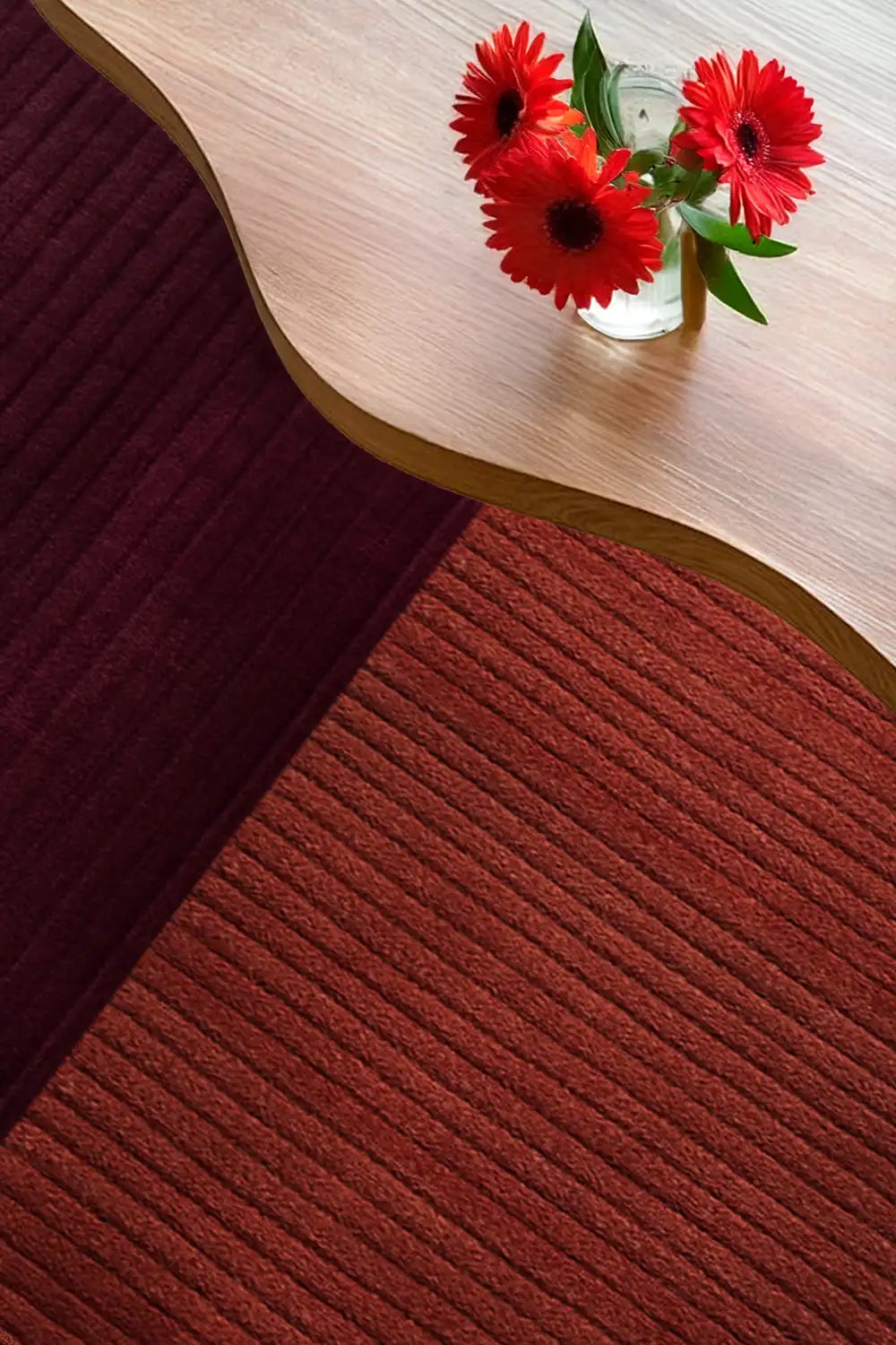Red flowers in a glass vase on a wooden surface with a textured red runner rug below.