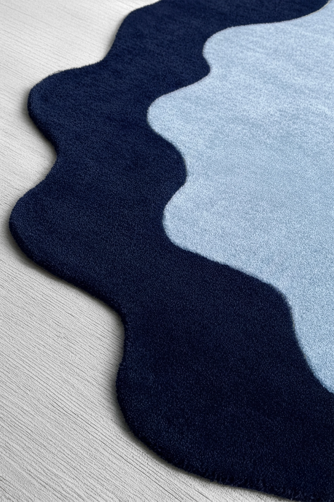 A close-up of a plush area rug with a wavy scalloped edge, featuring a dark navy blue outer section and a light blue inner section on a light gray wood floor.