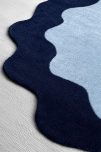 A close-up of a plush area rug with a wavy scalloped edge, featuring a dark navy blue outer section and a light blue inner section on a light gray wood floor.