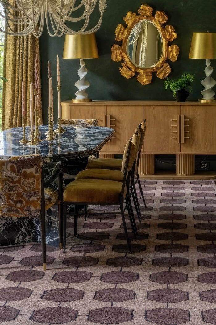 A styled interior scene featuring a patterned area rug with dark brown hexagonal motifs on a walnut base, a dining table with a blue-and-white marble top, upholstered chairs, a wooden sideboard, gold lamps, and a decorative mirror against a dark green wall.