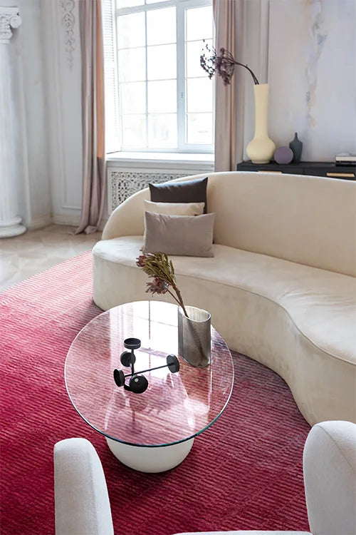 Modern living room with a curved beige sofa, glass coffee table, and pink wool rug.