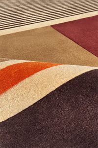Close-up of a textured area rug with a modern geometric pattern featuring wavy and angular color blocks in dark brown, rust orange, cream, tan, burgundy, and black.