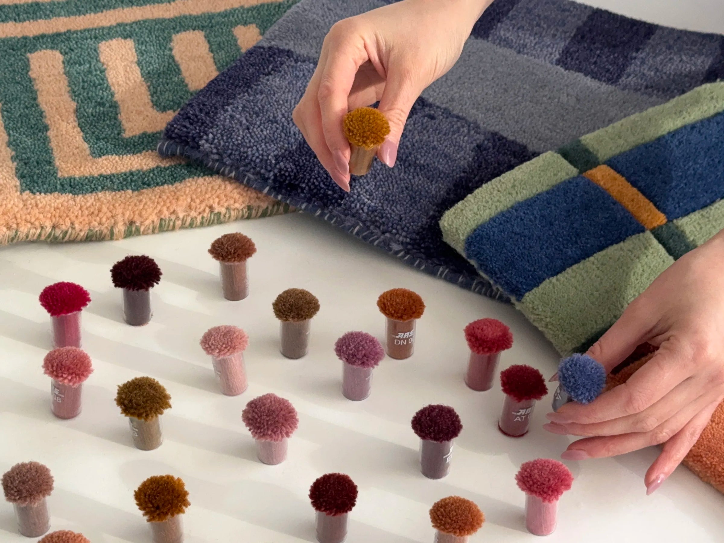 Person arranging small rug samples on a white surface with patterned swatches in the background.