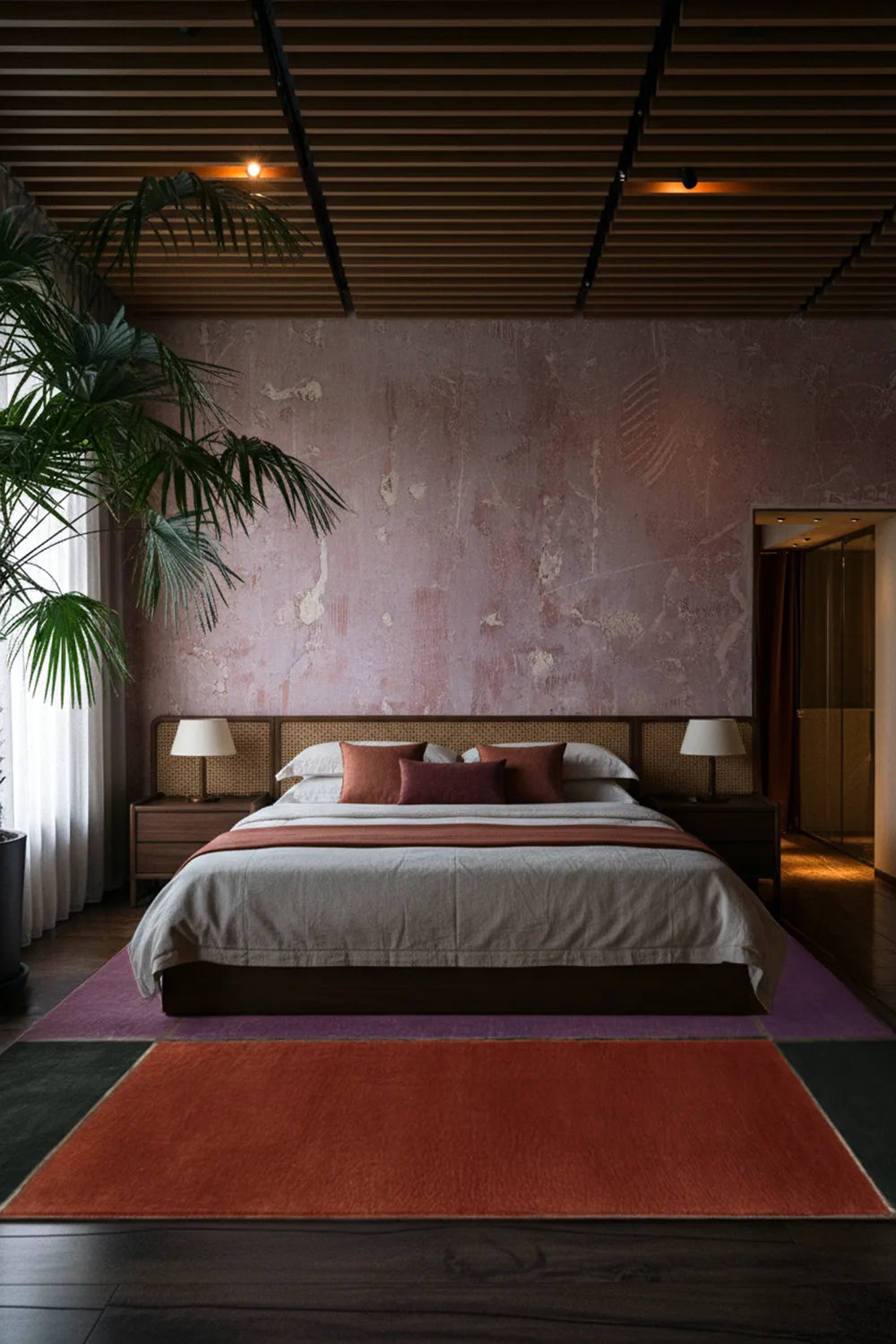 Modern bedroom with a geometric purple, orange and green wool rug with contemporary bed, lamps, and decorative pillows against a textured wall.